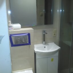 Norbert Stępień - Jasna łazienka po remoncie: biała umywalka z szafką, toaleta i lustro. Widoczna kabina prysznicowa. Zabezpieczony taśmą przycisk spłuczki.