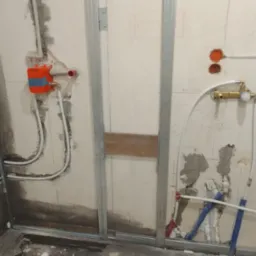 Ściana w trakcie montażu instalacji wodno-kanalizacyjnej z widocznymi rurami, stelażami metalowymi i osadzonymi elementami armatury w surowym stanie.
