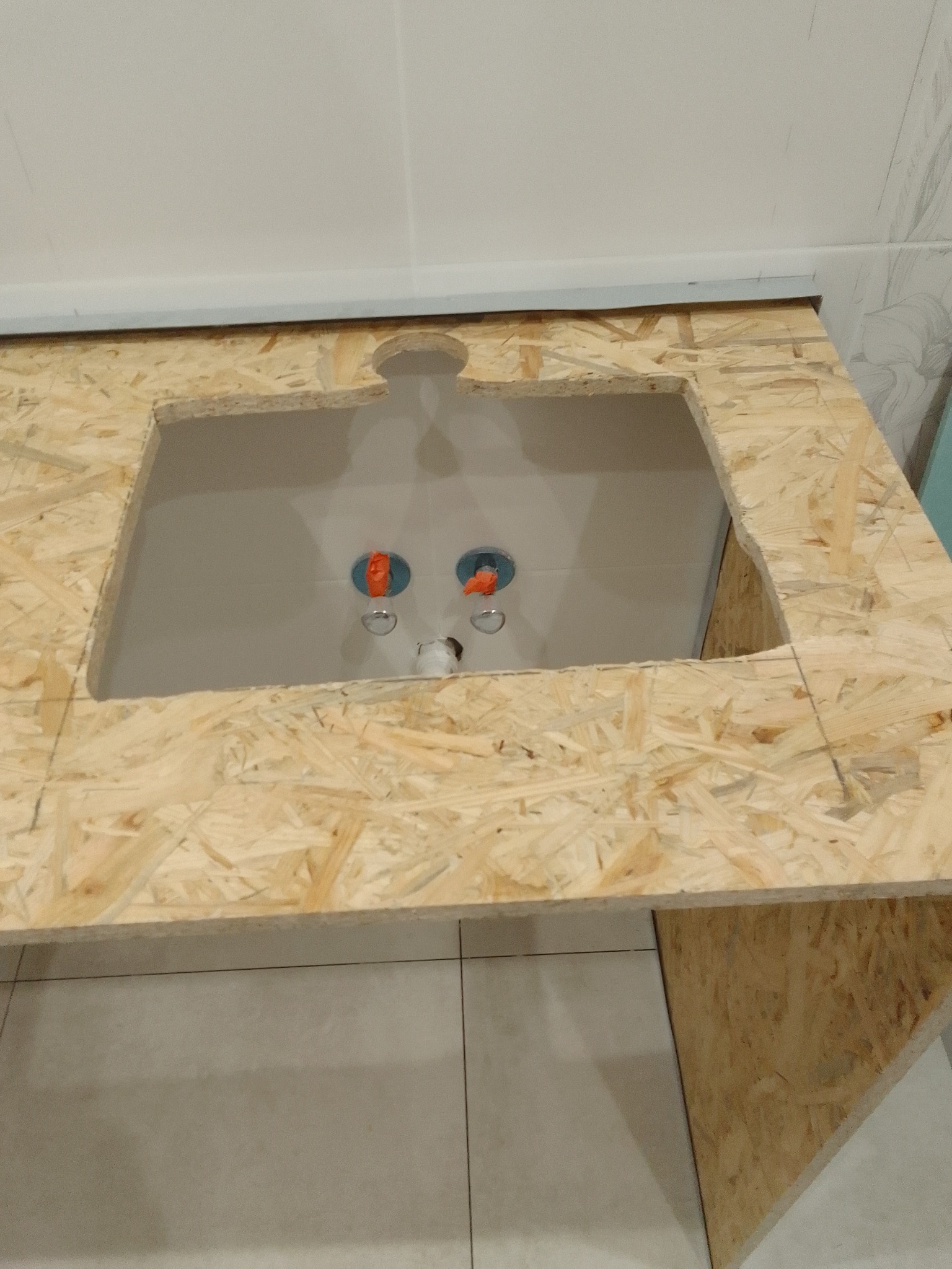 Szafka umywalkowa z płyty OSB w trakcie montażu, widoczne przyłącza wodne. Surowy stan wykończenia blatu i podłogi z płytek.