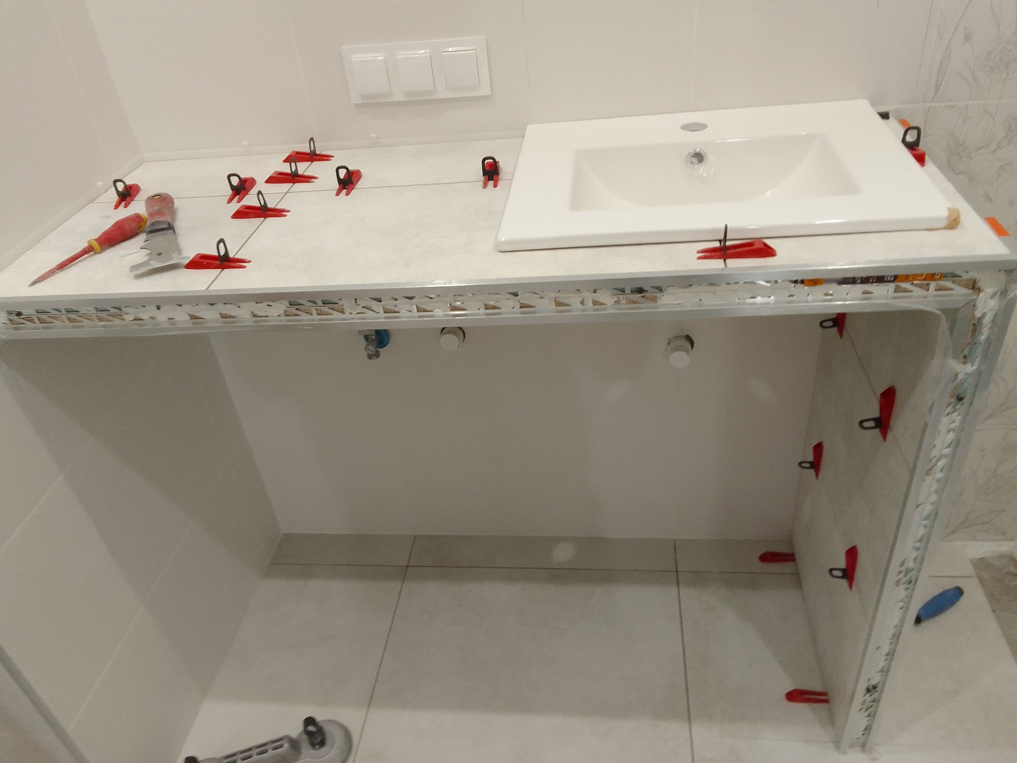 Biała umywalka osadzona na blacie z jasnych płytek, zabezpieczonych czerwonymi klipsami. W tle białe ściany i podłoga w trakcie prac wykończeniowych w łazience.