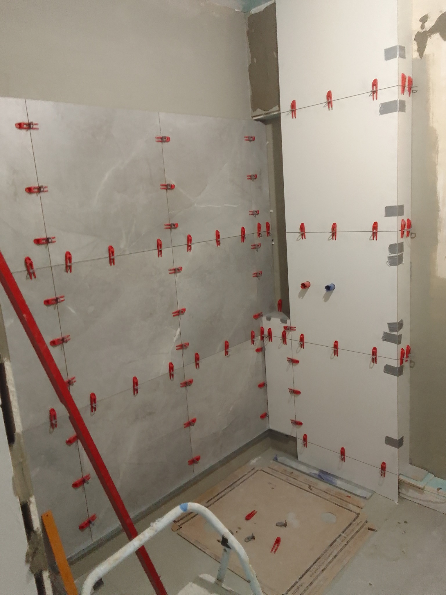 Układanie płytek w łazience. Widoczne płytki na ścianach z czerwonymi klipsami poziomującymi, poziomica i fragment brodzika. Prace w toku.