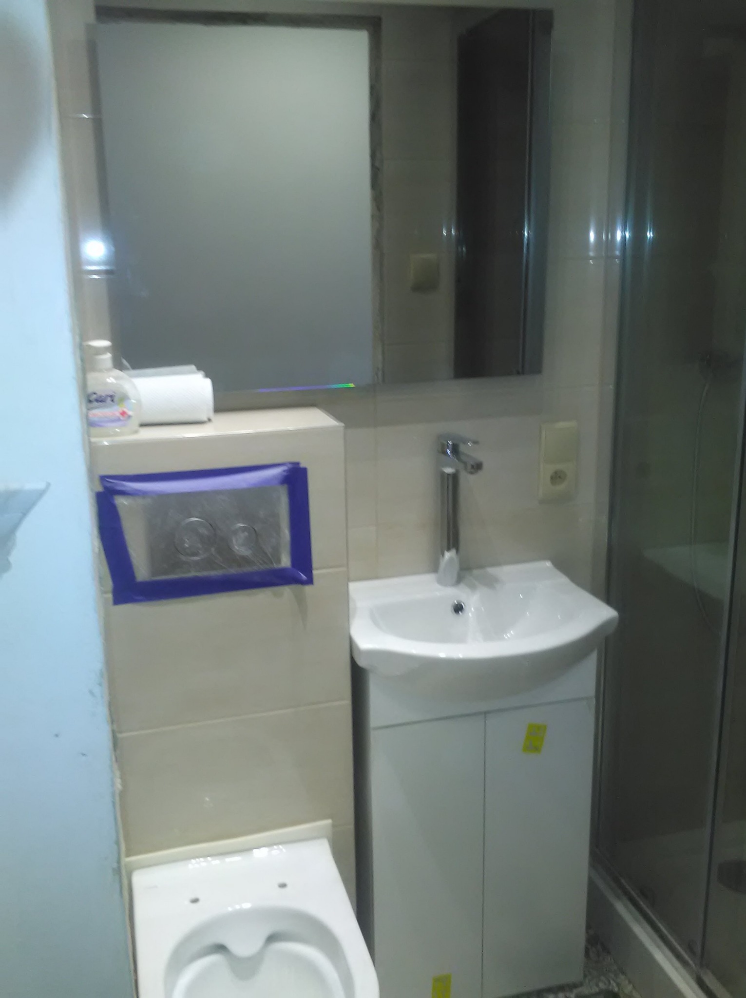 Jasna łazienka po remoncie: biała umywalka z szafką, toaleta i lustro. Widoczna kabina prysznicowa. Zabezpieczony taśmą przycisk spłuczki.