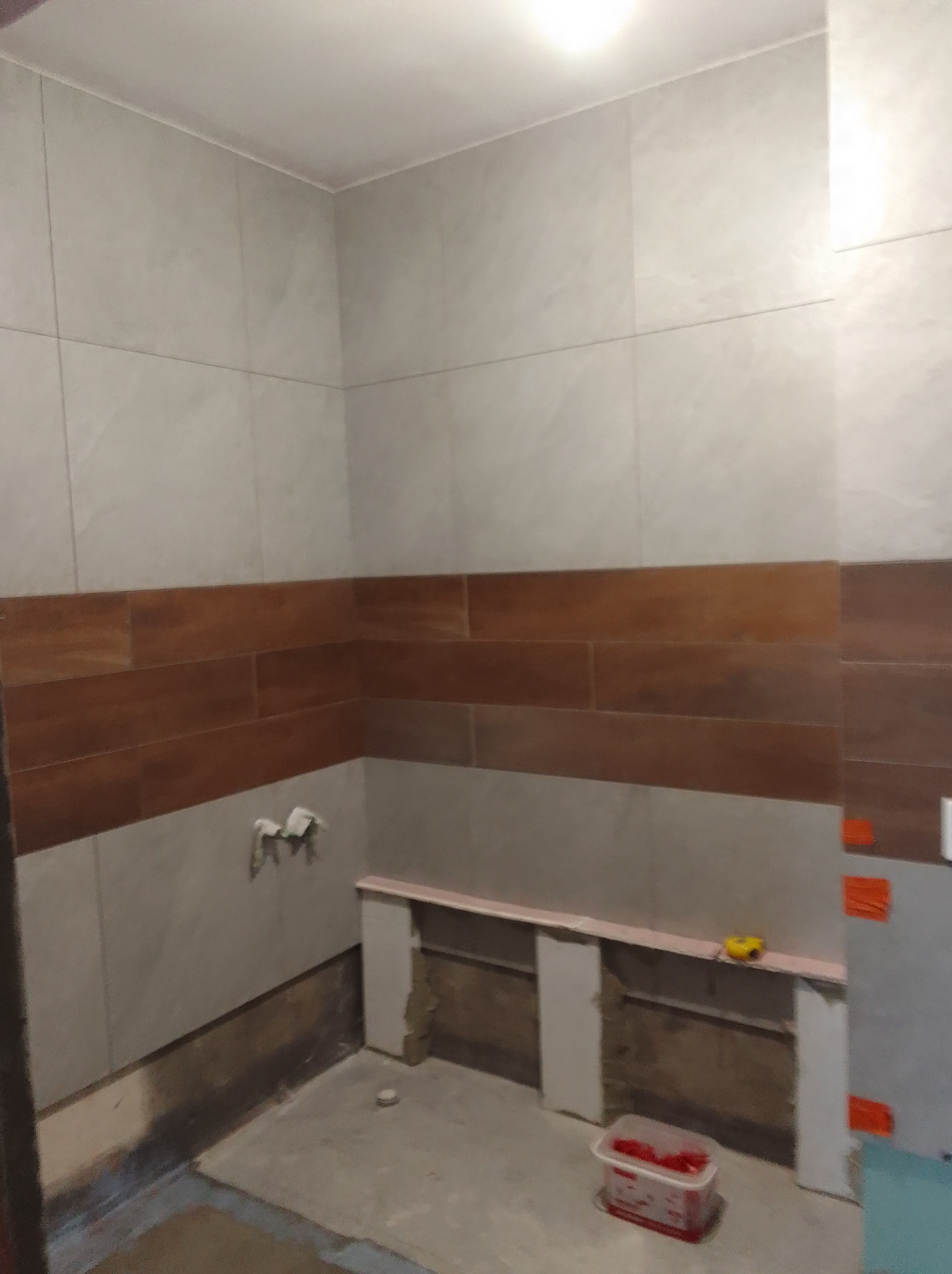 Łazienka w trakcie prac wykończeniowych: ułożone płytki ścienne w odcieniach szarości i brązu, widoczne narzędzia, przygotowane podłoże pod dalsze prace.