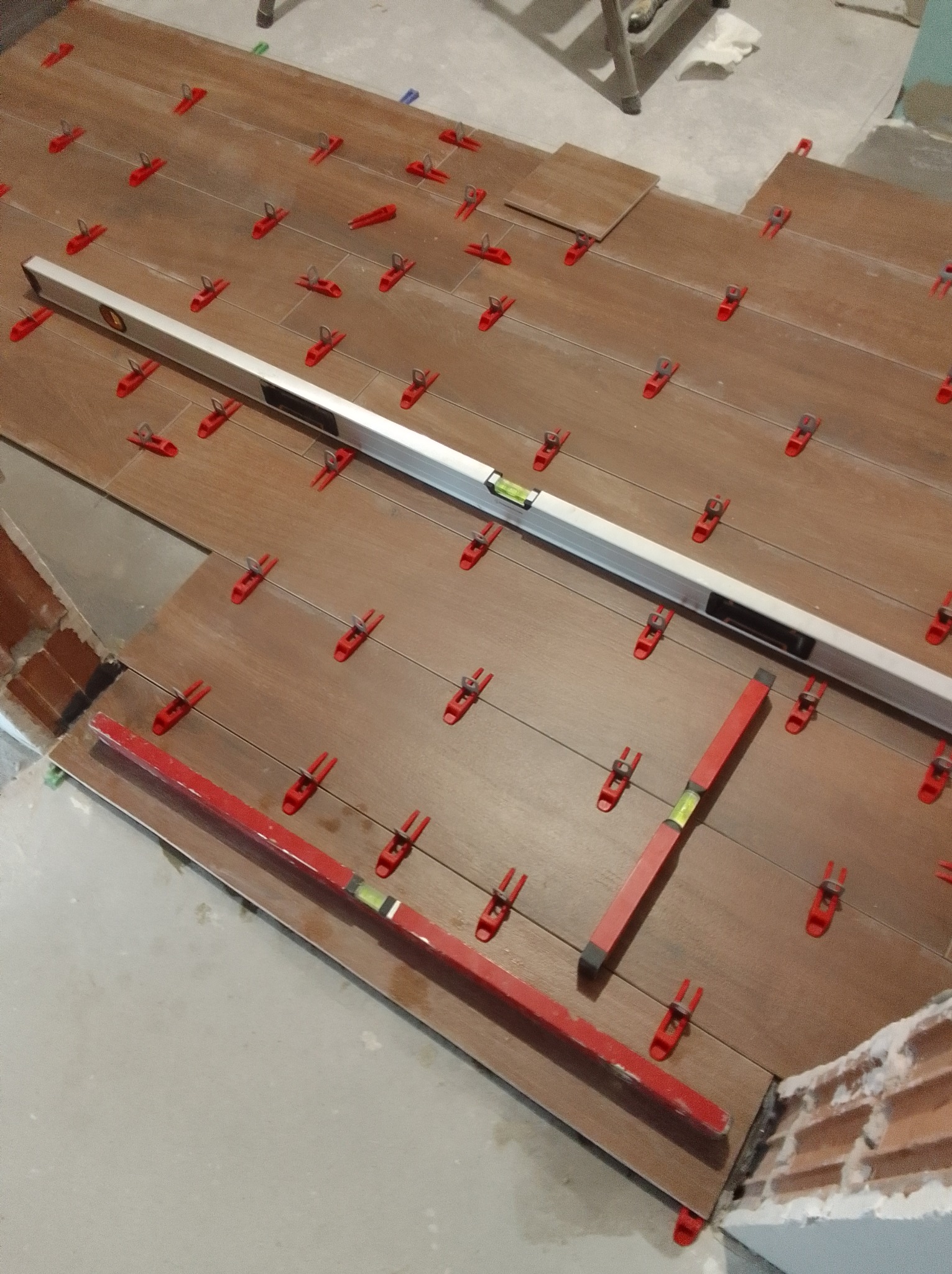 Układanie płytek imitujących drewno, poziomowanie z użyciem poziomicy i klipsów. Widoczne fragmenty ściany z cegły i podłogi.