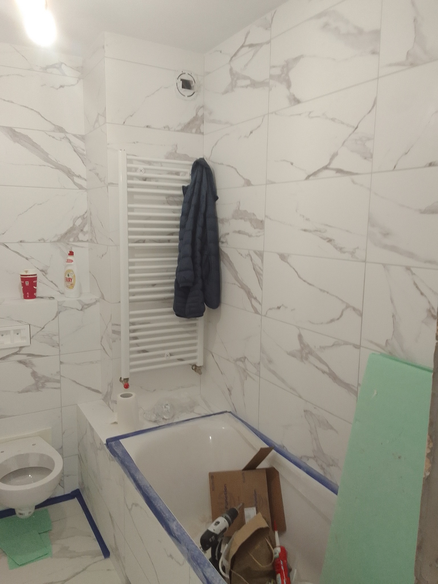 Remont łazienki w trakcie: białe płytki imitujące marmur, wanna z niebieskim obramowaniem, wiszący grzejnik i narzędzia w wannie.