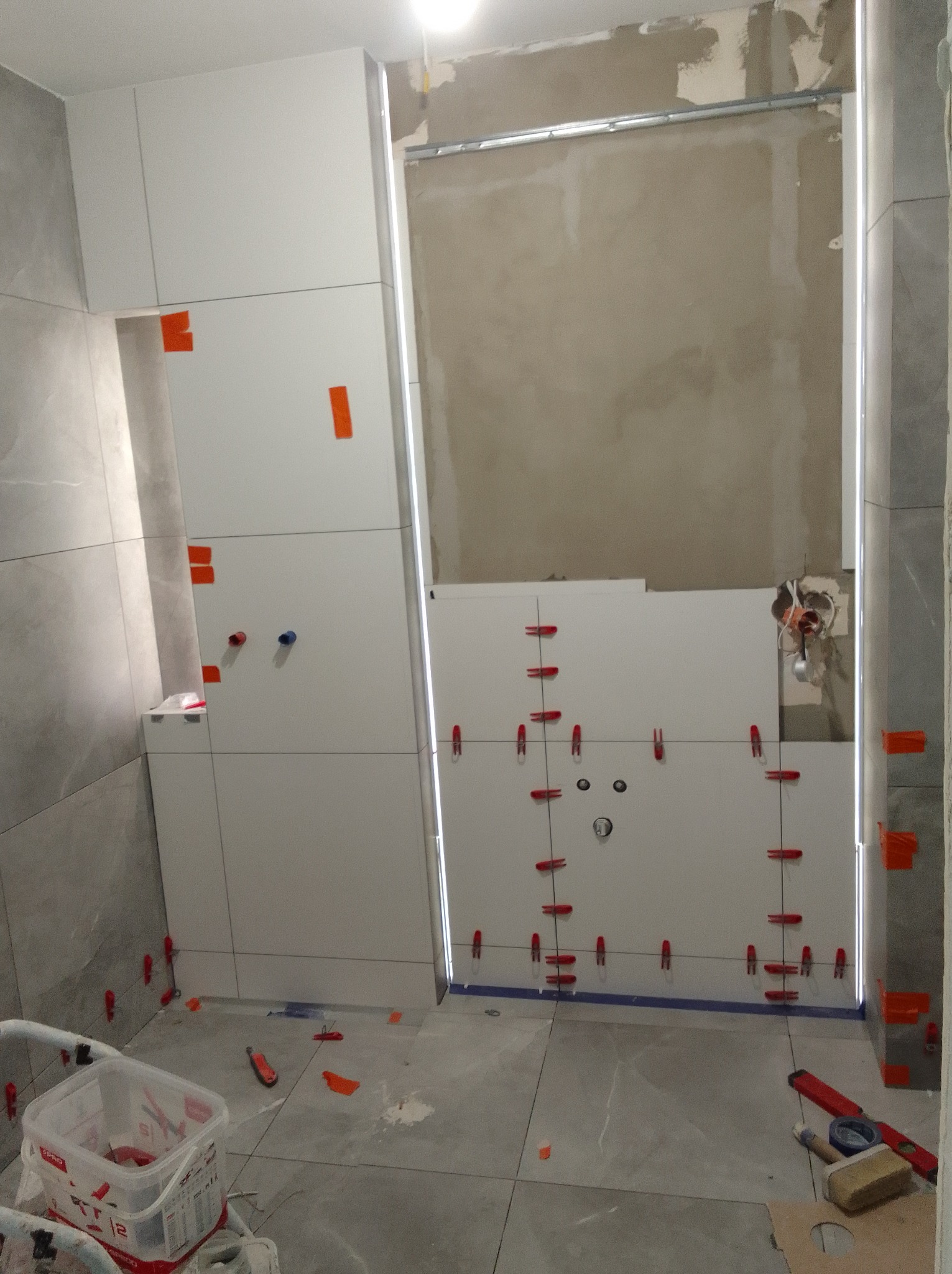 Układanie białych płytek w łazience z użyciem krzyżyków dystansowych. Widoczne narzędzia glazurnicze i fragmenty niedokończonej ściany. Szare płytki na podłodze i ścianie.