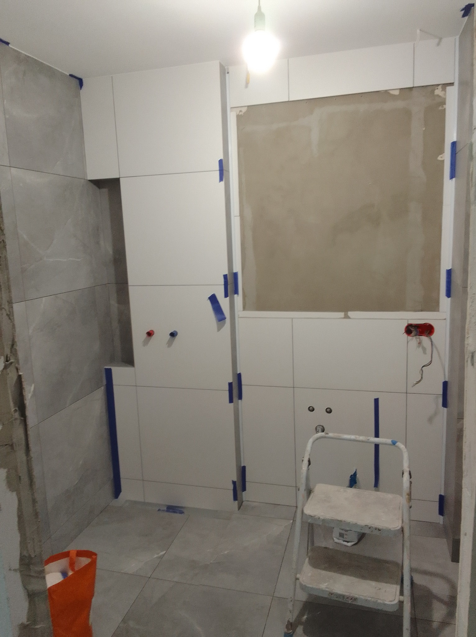 Łazienka w trakcie wykańczania: szare płytki na ścianie, białe płytki na ścianie z wyciętym miejscem, drabina malarska, zabezpieczenia z taśmy malarskiej.