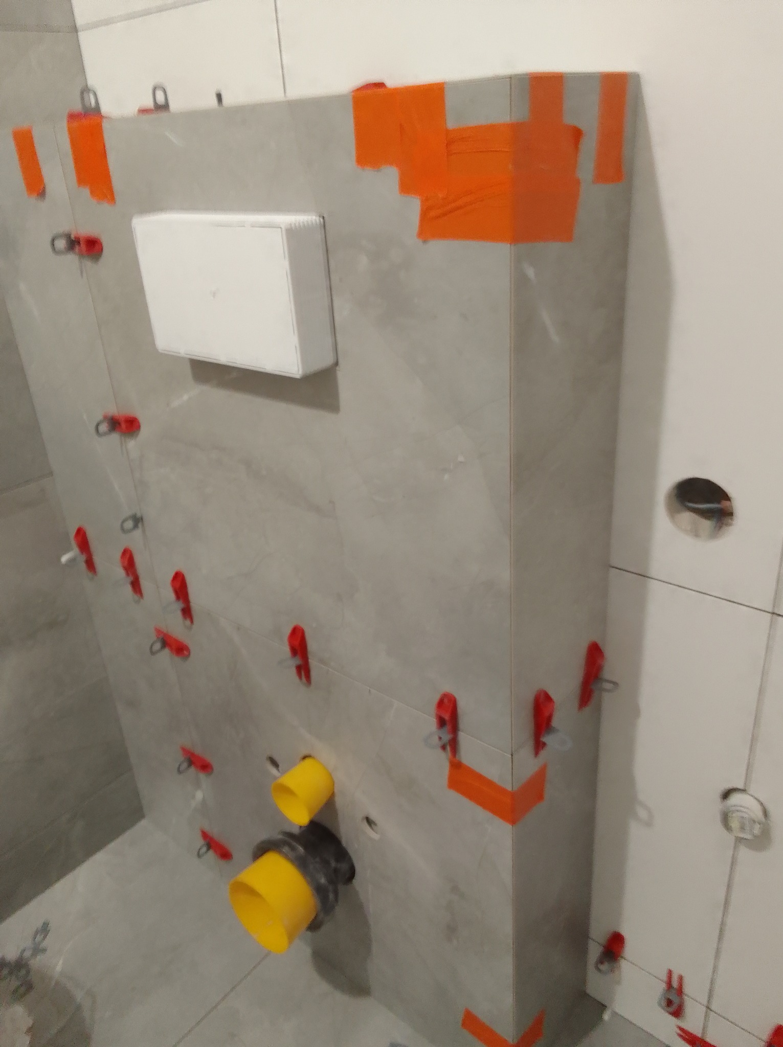 Szare płytki imitujące kamień, zamontowane z użyciem klipsów i taśmy, tworzą obudowę stelaża WC. Widoczne żółte rury i otwory na instalacje.
