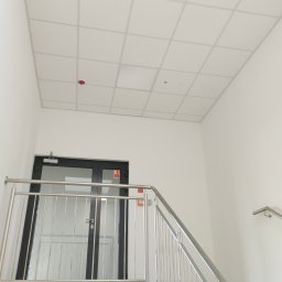Vitos - Biały sufit kasetonowy w jasnym pomieszczeniu biurowym, widoczny fragment schodów z metalową barierką i drzwi ewakuacyjne. Czerwony czujnik dymu na suficie.