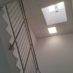 Vitos - Widok z dołu na klatkę schodową z metalową balustradą. Na suficie widoczne kwadratowe panele i okno dachowe z drabiną. Minimalistyczne wykończenie wnętrza.