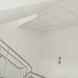 Vitos - Białe wnętrze klatki schodowej z metalową balustradą i sufitem kasetonowym. Na ścianie widoczny pomarańczowy panel alarmowy.