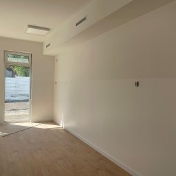 Vitos - Jasne wnętrze po malowaniu z drewnopodobną podłogą, widoczne gniazdka elektryczne i listwy przypodłogowe, okno z widokiem na zewnątrz, minimalistyczne oświetlenie sufitowe.