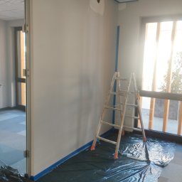 Vitos - Przygotowania do malowania ścian w biurze: drabina, folia ochronna na podłodze i taśma malarska przy suficie i oknach. Ściany w odcieniach szarości, widoczne ślady po szpachlowaniu.