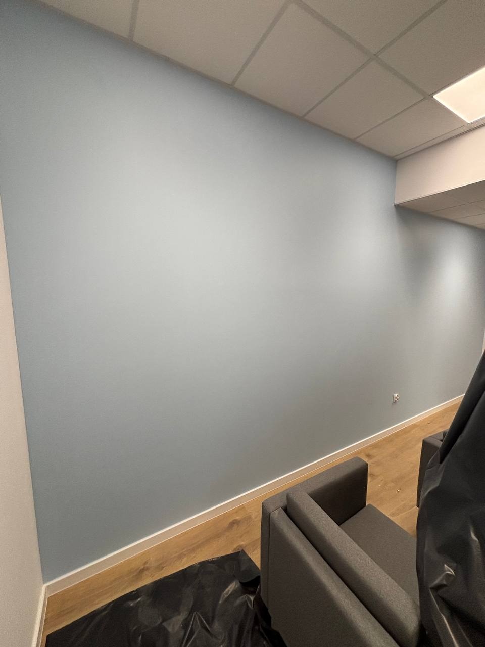 Ściana pomalowana na jasny, pastelowy odcień niebieskiego w biurze, z widocznym zabezpieczeniem podłogi i mebli. Świeżo po malowaniu.