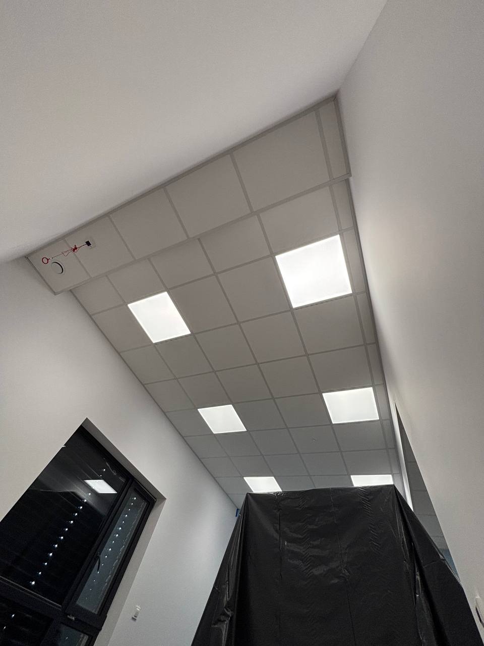 Sufit podwieszany kasetonowy z wbudowanym oświetleniem LED, widok z dołu, fragment okna i zabezpieczone folią miejsce pracy.