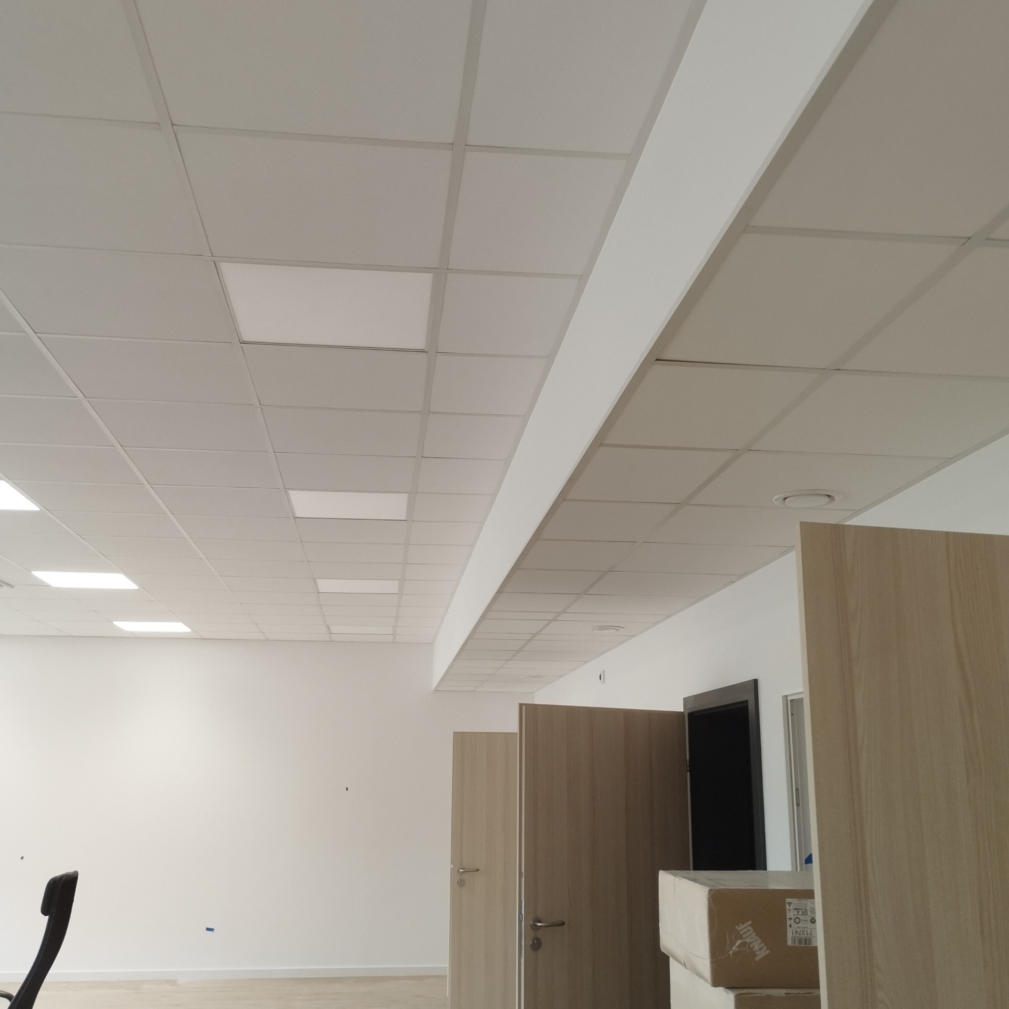 Biały sufit kasetonowy z panelami LED w nowym biurze. Widoczne drzwi wewnętrzne i kartony, co sugeruje wykańczanie wnętrz. Minimalistyczny design.