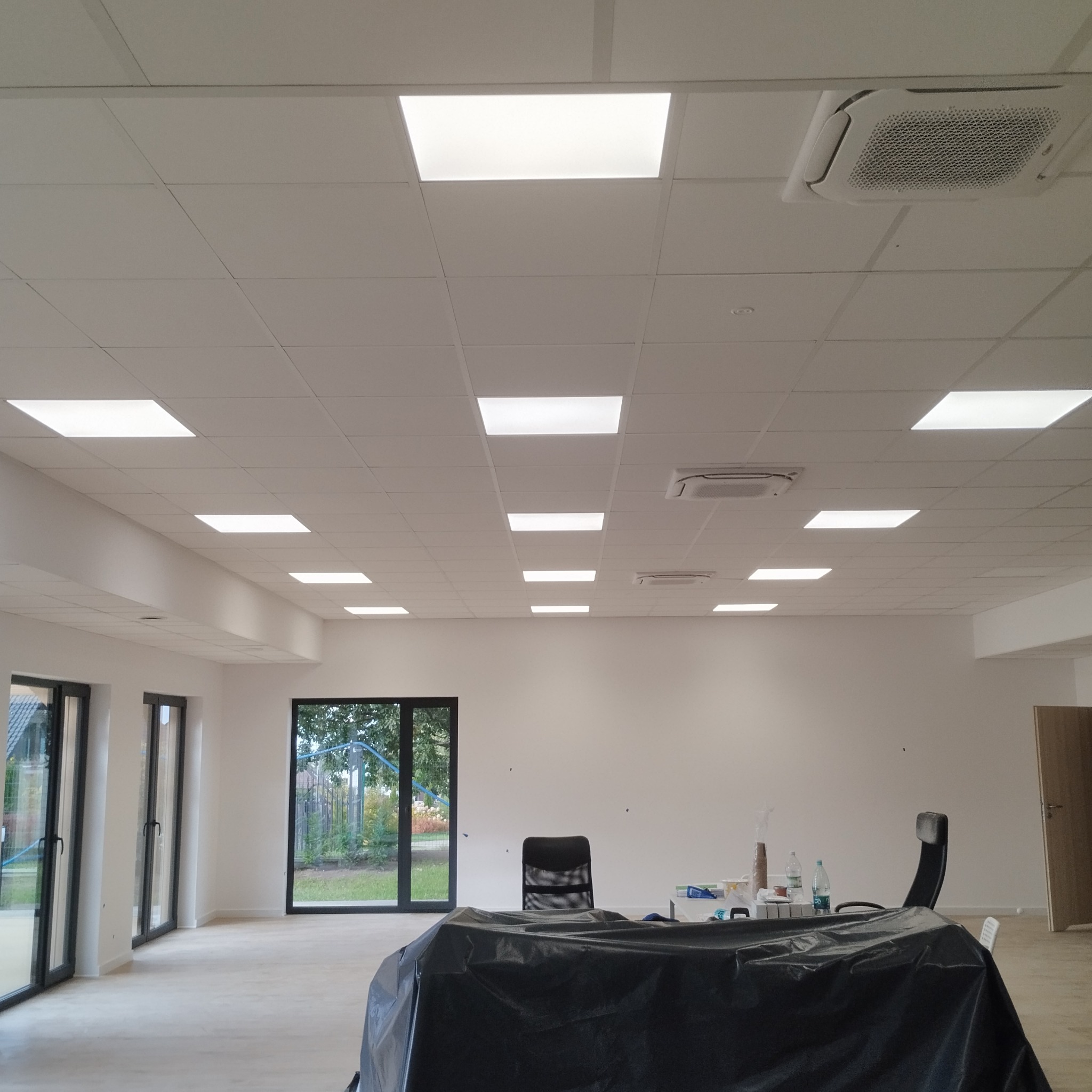 Białe, kasetonowe sufity z wbudowanym oświetleniem w jasnym pomieszczeniu biurowym. Widoczne klimatyzatory, okna i meble zakryte folią ochronną.