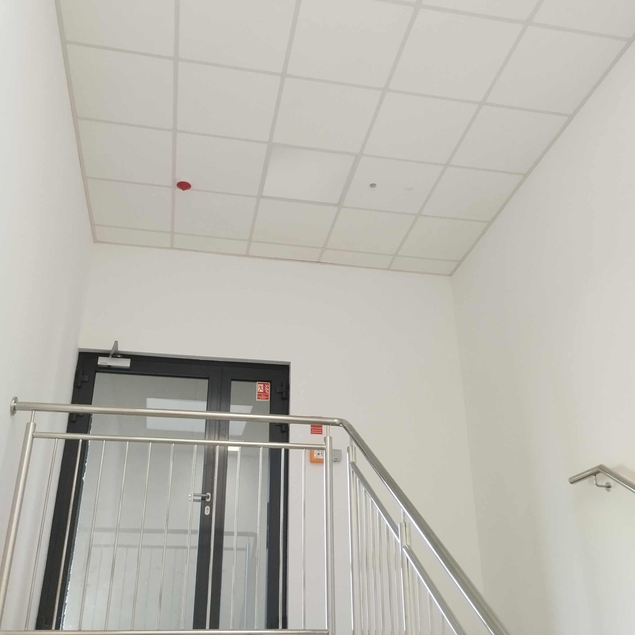 Biały sufit kasetonowy w jasnym pomieszczeniu biurowym, widoczny fragment schodów z metalową barierką i drzwi ewakuacyjne. Czerwony czujnik dymu na suficie.