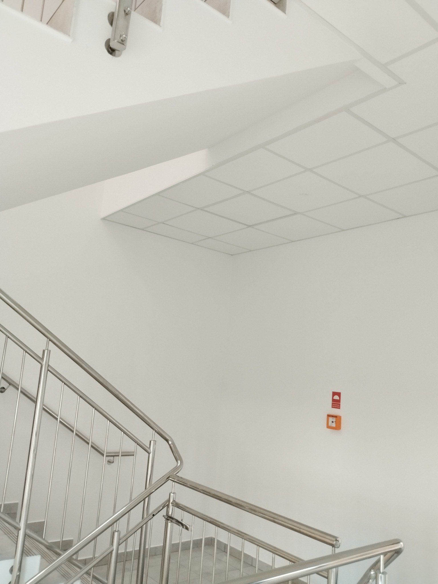 Białe wnętrze klatki schodowej z metalową balustradą i sufitem kasetonowym. Na ścianie widoczny pomarańczowy panel alarmowy.