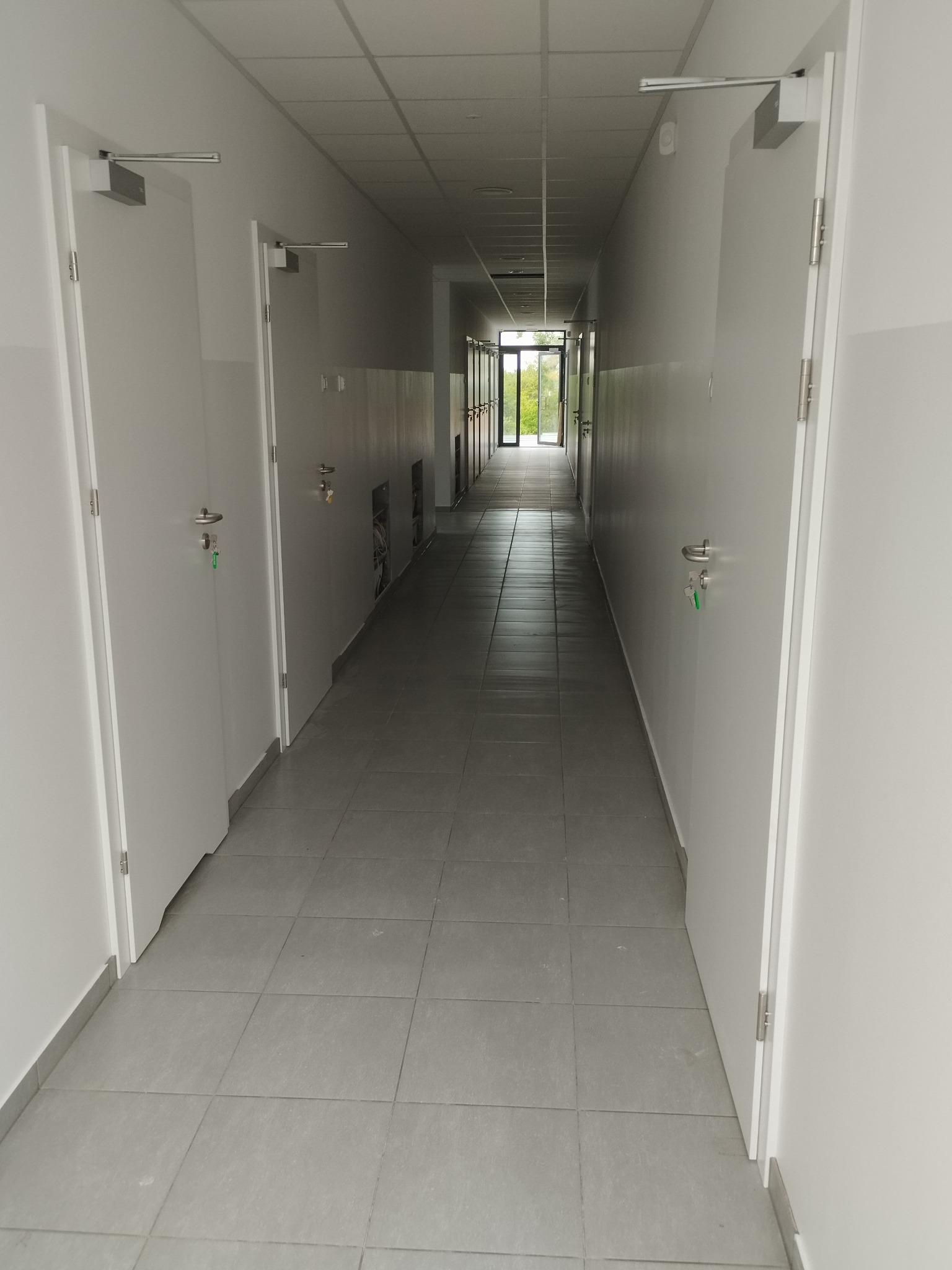 Długi, jasny korytarz biurowy z białymi drzwiami po obu stronach i szarą podłogą z płytek. Widoczne metalowe okucia drzwi i otwory wentylacyjne w ścianach.