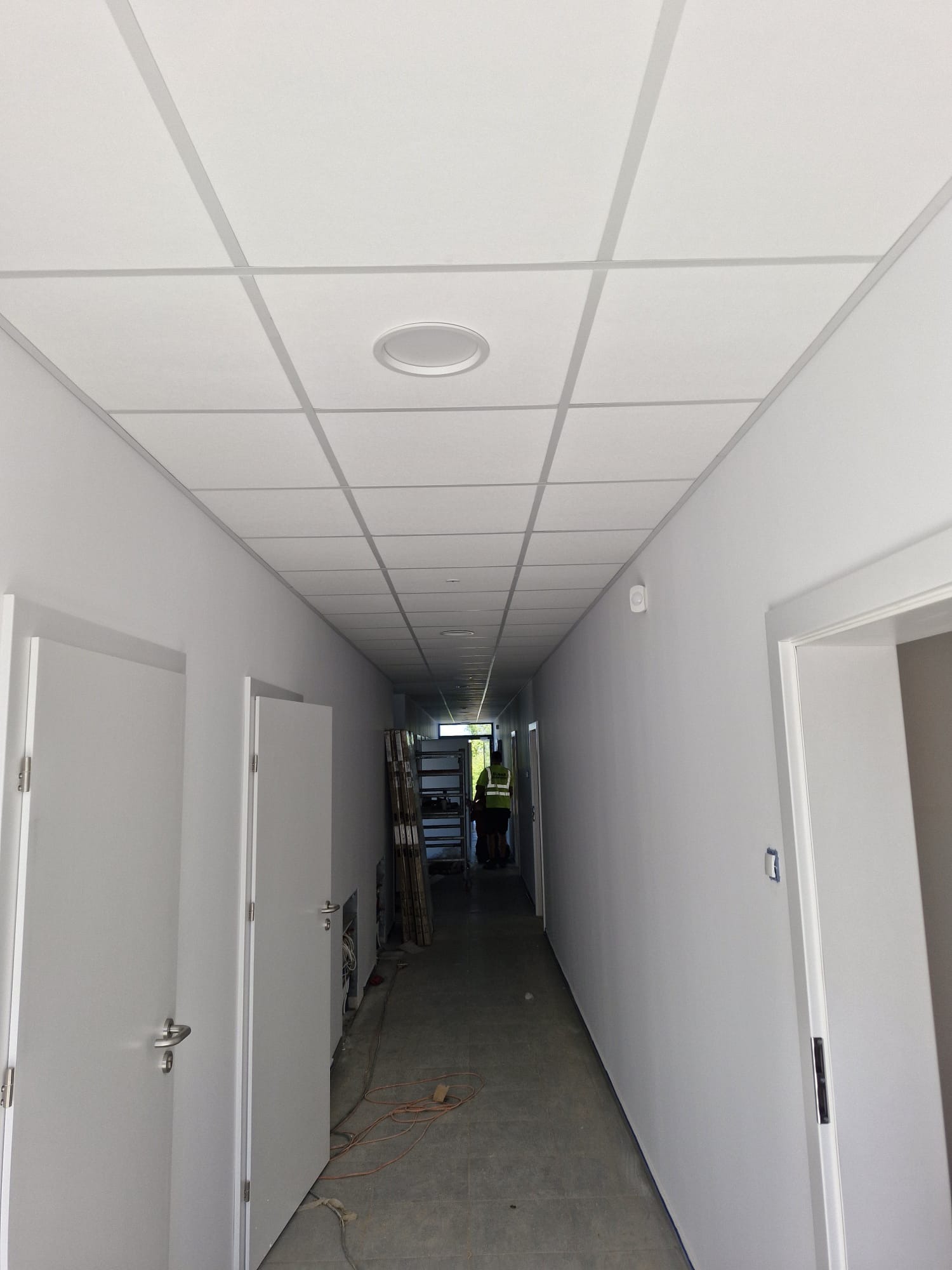 Korytarz biurowy z podwieszanym sufitem kasetonowym z oświetleniem, świeżo po remoncie. Widoczne białe ściany i drzwi, prace wykończeniowe w toku.