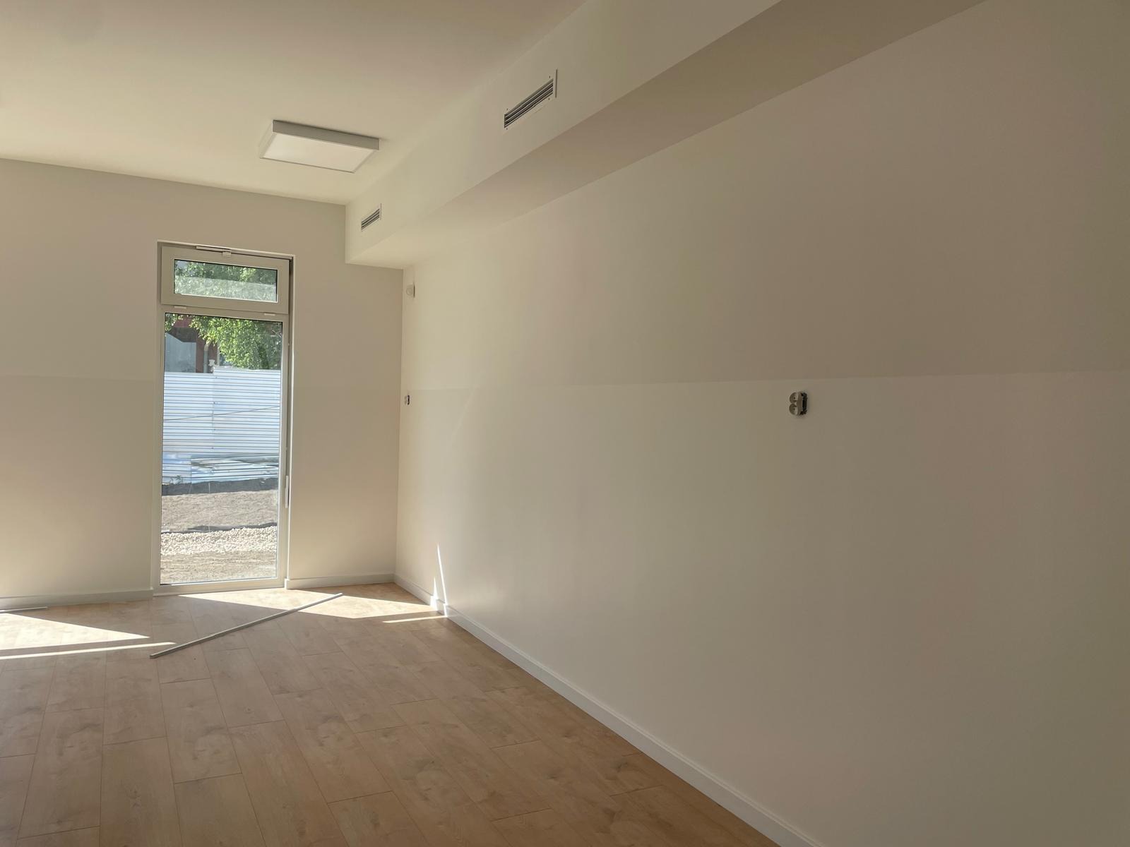 Jasne wnętrze po malowaniu z drewnopodobną podłogą, widoczne gniazdka elektryczne i listwy przypodłogowe, okno z widokiem na zewnątrz, minimalistyczne oświetlenie sufitowe.