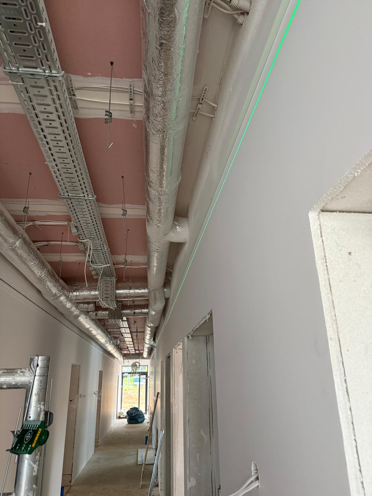 Korytarz w trakcie prac wykończeniowych z widocznymi instalacjami pod sufitem i laserowo wyznaczonym poziomem na świeżo pomalowanej ścianie.
