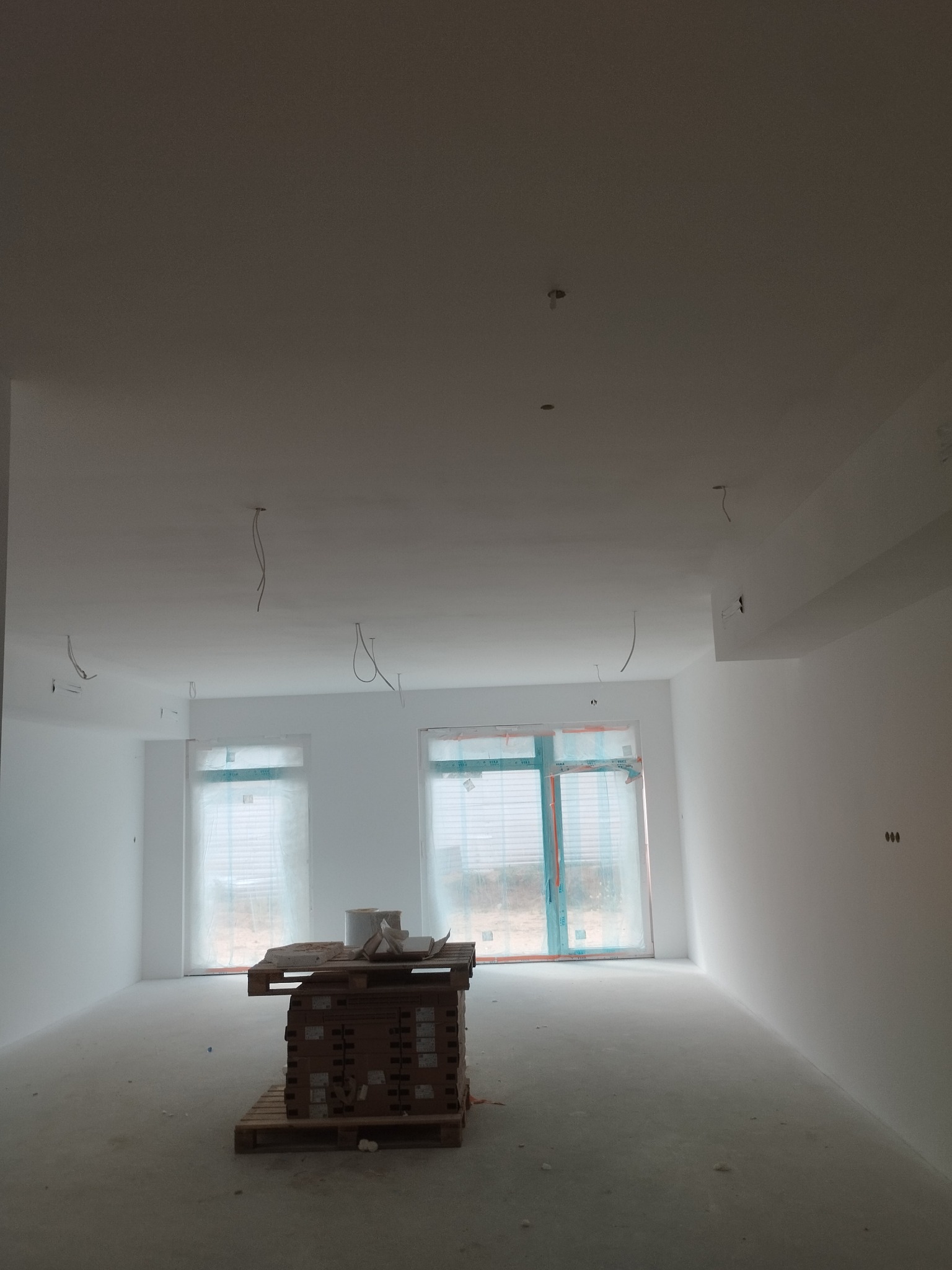 Białe, świeżo wyszpachlowane wnętrze pokoju z paletą materiałów budowlanych. Widoczne kable zwisające z sufitu i okna zabezpieczone folią.