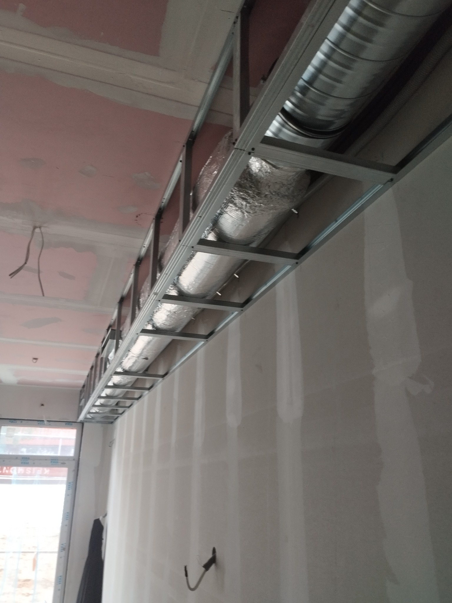 Montaż konstrukcji sufitu podwieszanego z widocznymi przewodami wentylacyjnymi. Ściany przygotowane do gładzi. Widok na okno w tle.