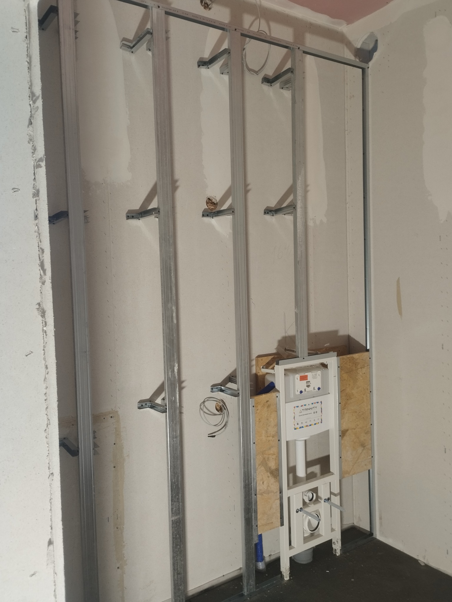 Montaż stelaża WC na ścianie z profilem pionowym; widoczne mocowania, instalacja i płyta OSB. Ściana przygotowana do obłożenia płytą gipsową.
