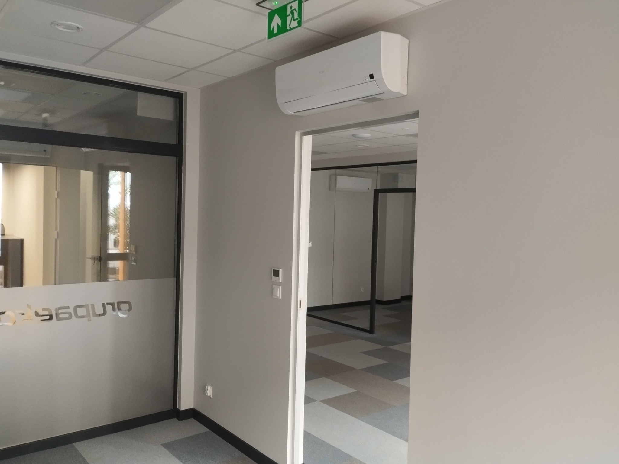 Jasne, minimalistyczne wnętrze biurowe z klimatyzacją, szklaną ścianą z logo i wzorzystą podłogą. Widoczny znak ewakuacyjny.