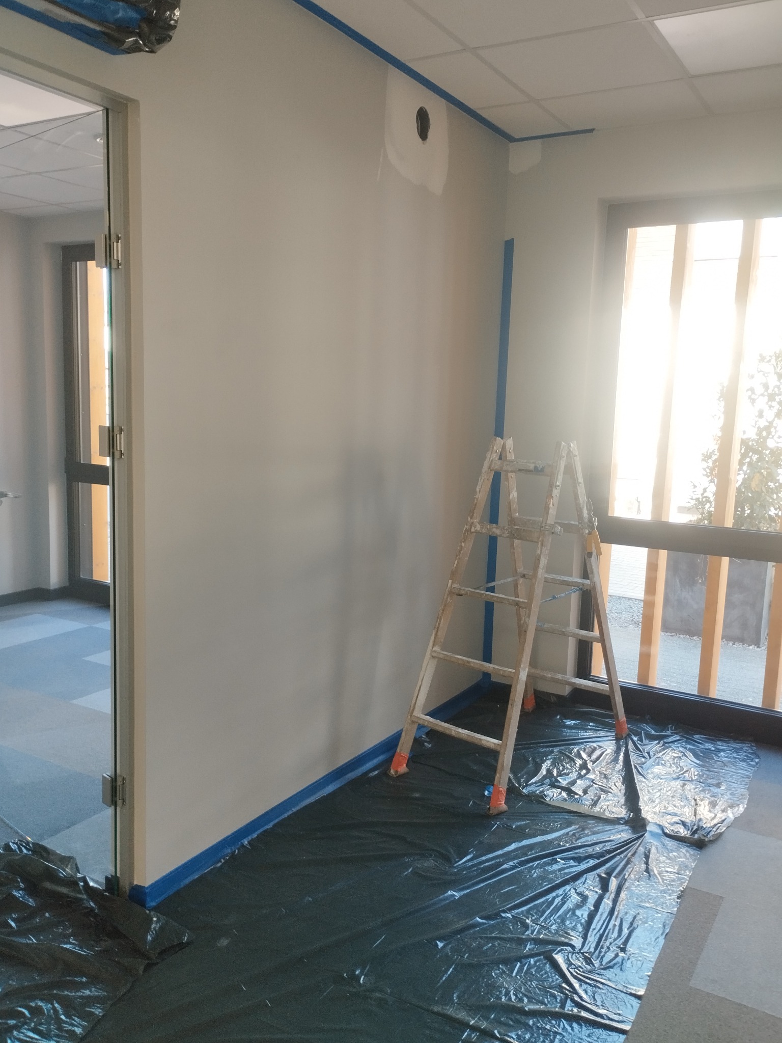 Przygotowania do malowania ścian w biurze: drabina, folia ochronna na podłodze i taśma malarska przy suficie i oknach. Ściany w odcieniach szarości, widoczne ślady po szpachlowaniu.