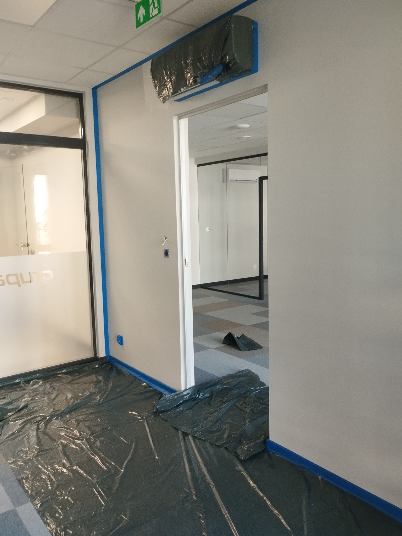 Bielone ściany biura zabezpieczone folią i taśmą malarską, klimatyzator zakryty, przygotowania do malowania.