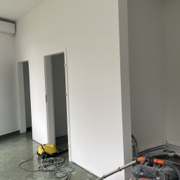 APF DECOR PAWEŁ SOCHACKI - Białe ściany po malowaniu w jasnym wnętrzu. Widoczne narzędzia malarskie i odkurzacz przemysłowy na zabrudzonej podłodze. Dwa wejścia bez drzwi.