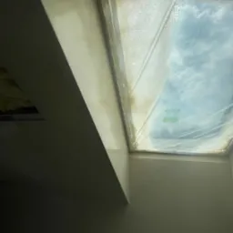 Pomalowane na gładko ściany wokół okna dachowego, zabezpieczonego folią ochronną. Widoczny fragment sufitu i narożnik pomieszczenia po remoncie.