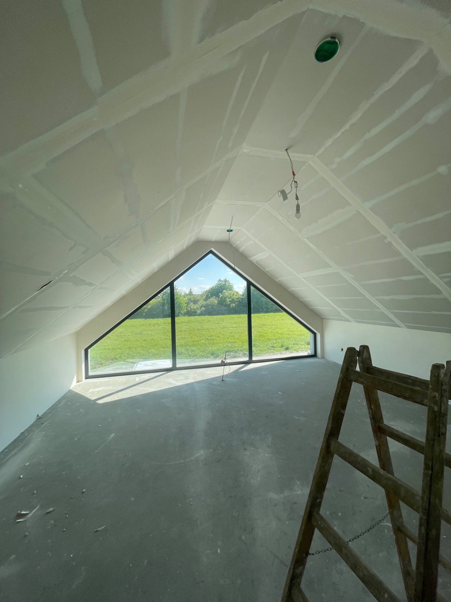 Poddasze z oknem w kształcie trójkąta, widok na zielone pole. Sufit i ściany pokryte płytami karton-gips, widoczne łączenia. Na pierwszym planie drewniana drabina.