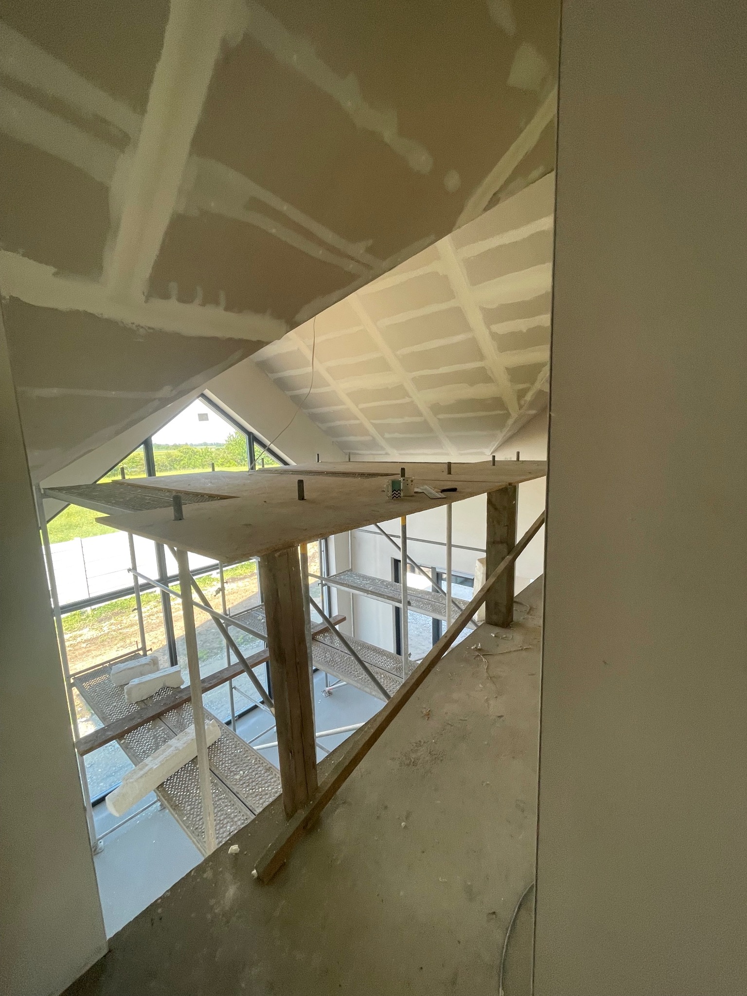 Skośny sufit z płyt kartonowo-gipsowych, częściowo zaszpachlowany, widok z góry na rusztowanie i okno. Prace wykończeniowe w nowym domu.