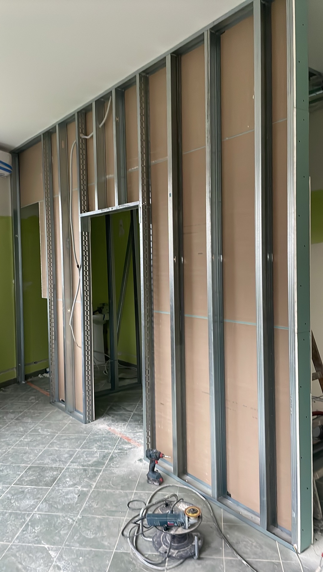 Konstrukcja ścian działowych z płyt gipsowo-kartonowych na metalowym stelażu, widoczne narzędzia: wiertarka i szlifierka kątowa na podłodze w remontowanym pomieszczeniu.