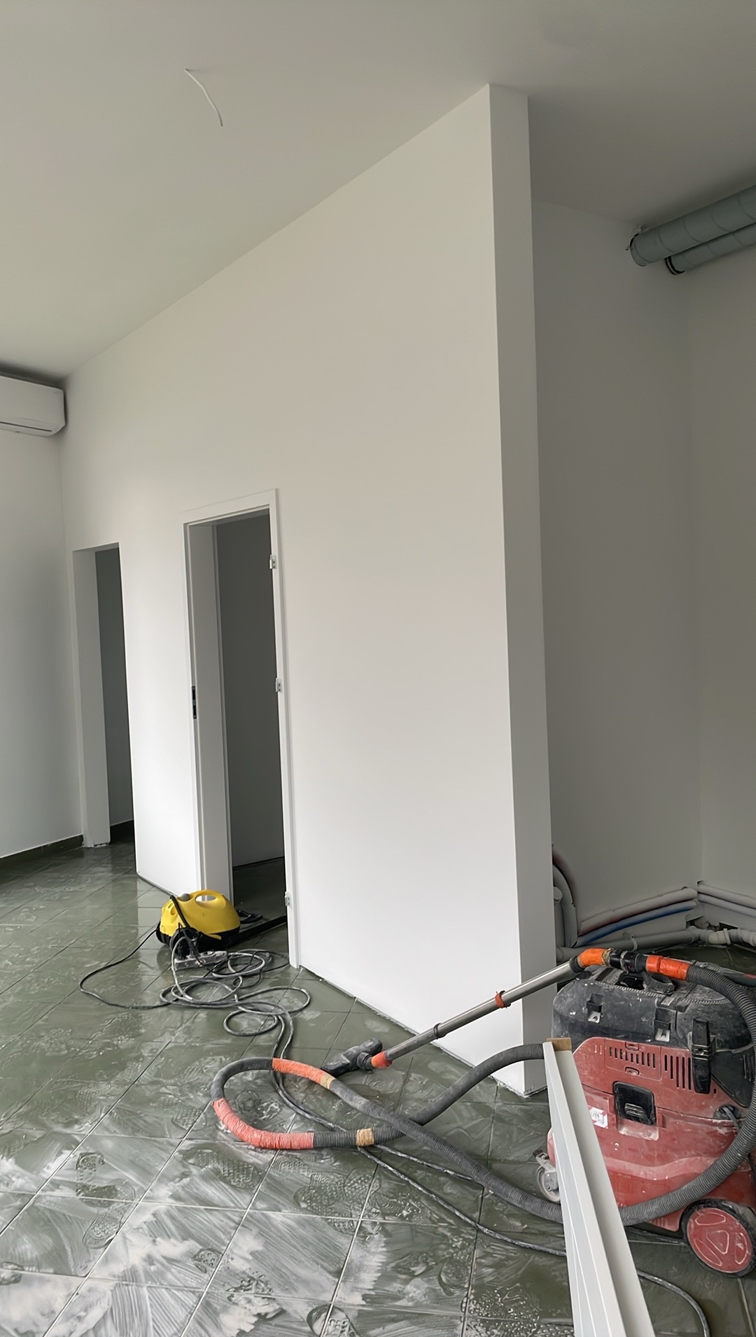 Białe ściany po malowaniu w jasnym wnętrzu. Widoczne narzędzia malarskie i odkurzacz przemysłowy na zabrudzonej podłodze. Dwa wejścia bez drzwi.