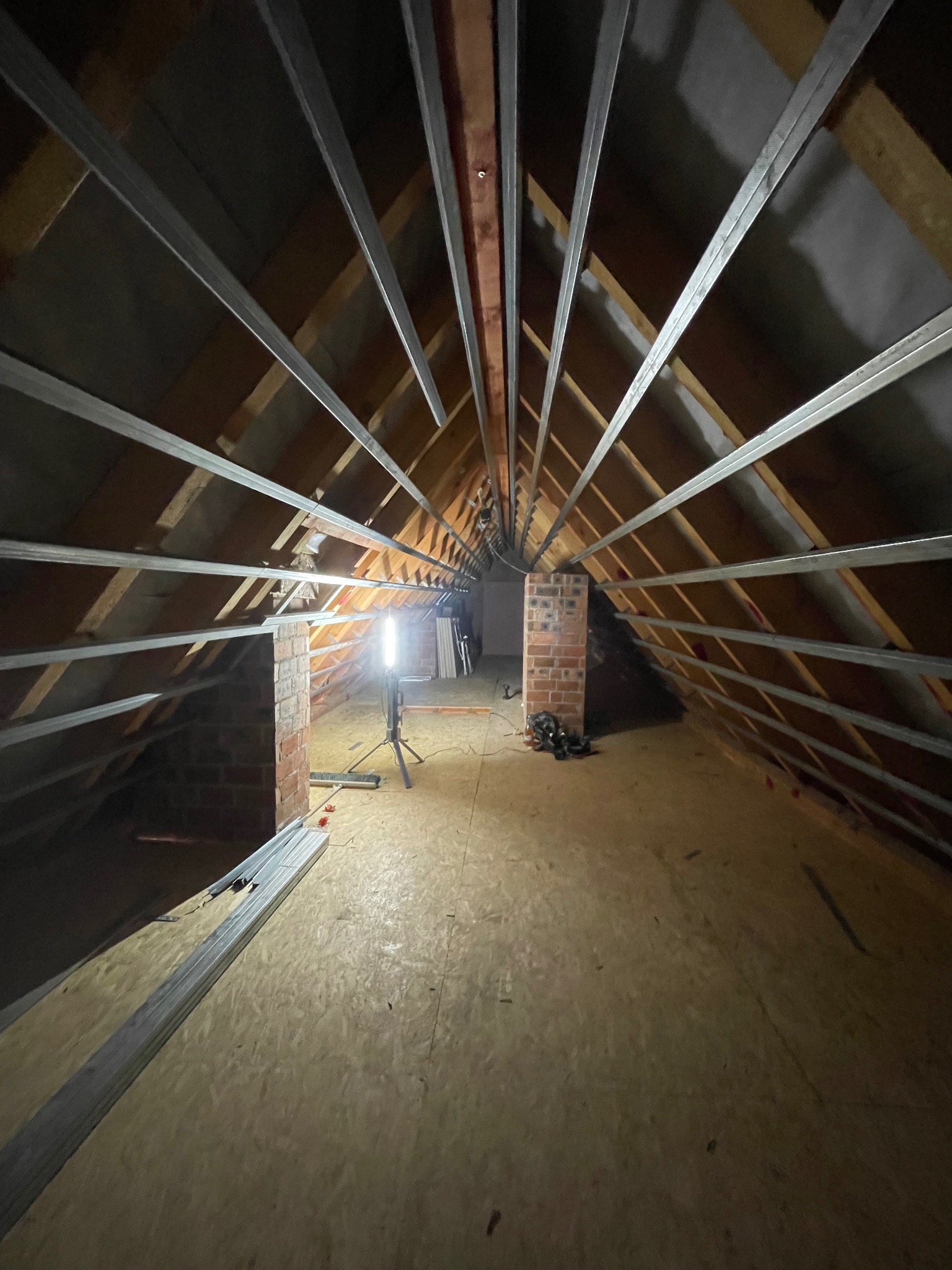 Montaż stelażu pod płyty G-K na poddaszu w Strzelinie. Widoczne profile metalowe, drewniana konstrukcja dachu i oświetlenie robocze. Prace wykończeniowe.