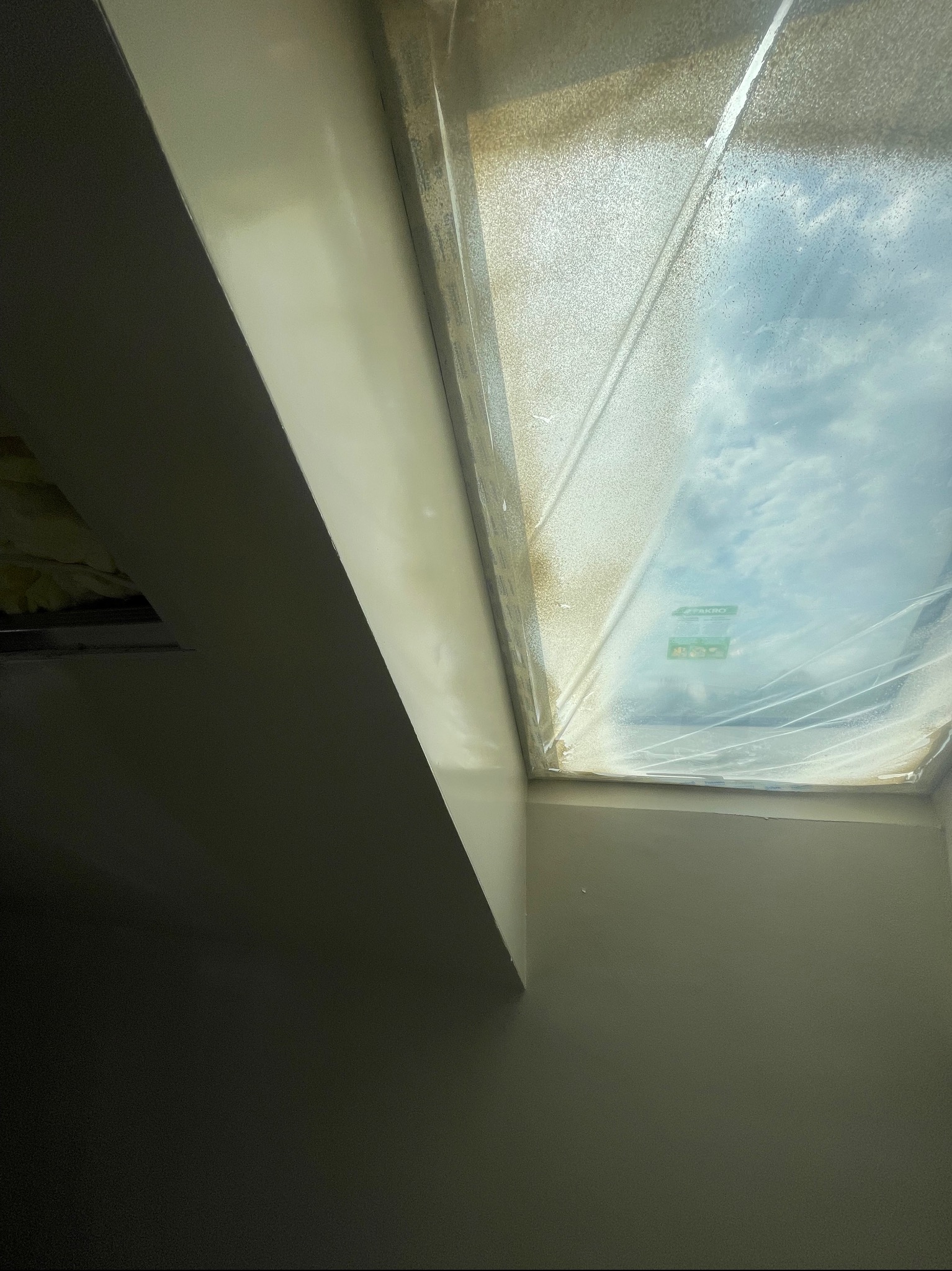 Pomalowane na gładko ściany wokół okna dachowego, zabezpieczonego folią ochronną. Widoczny fragment sufitu i narożnik pomieszczenia po remoncie.