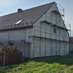 Dom w trakcie renowacji elewacji, otoczony rusztowaniami, z widocznym fragmentem ogrodu i sąsiednich budynków w słoneczny dzień. Widoczny surowy tynk na ścianach.