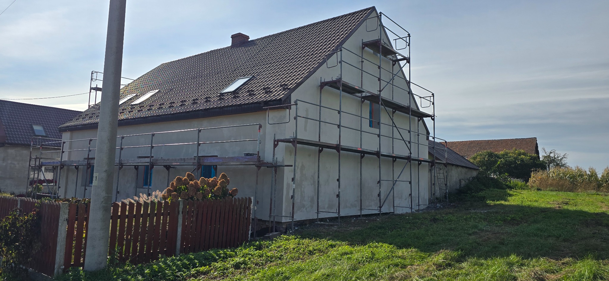 Dom w trakcie renowacji elewacji, otoczony rusztowaniami, z widocznym fragmentem ogrodu i sąsiednich budynków w słoneczny dzień. Widoczny surowy tynk na ścianach.