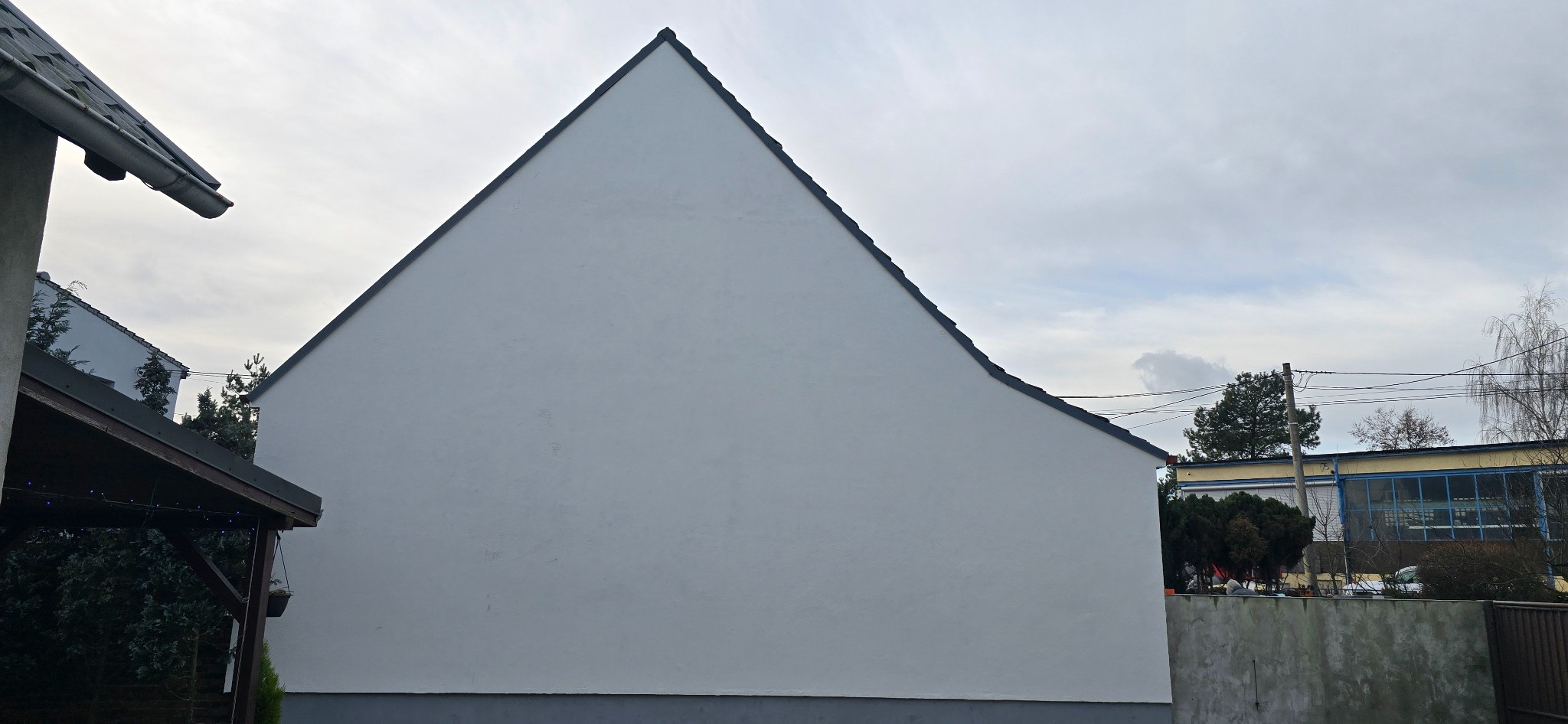 Świeżo otynkowana, biała ściana szczytowa domu jednorodzinnego, widoczna z boku, z fragmentem dachu i otoczenia. Minimalistyczny wygląd, pochmurne niebo w tle.