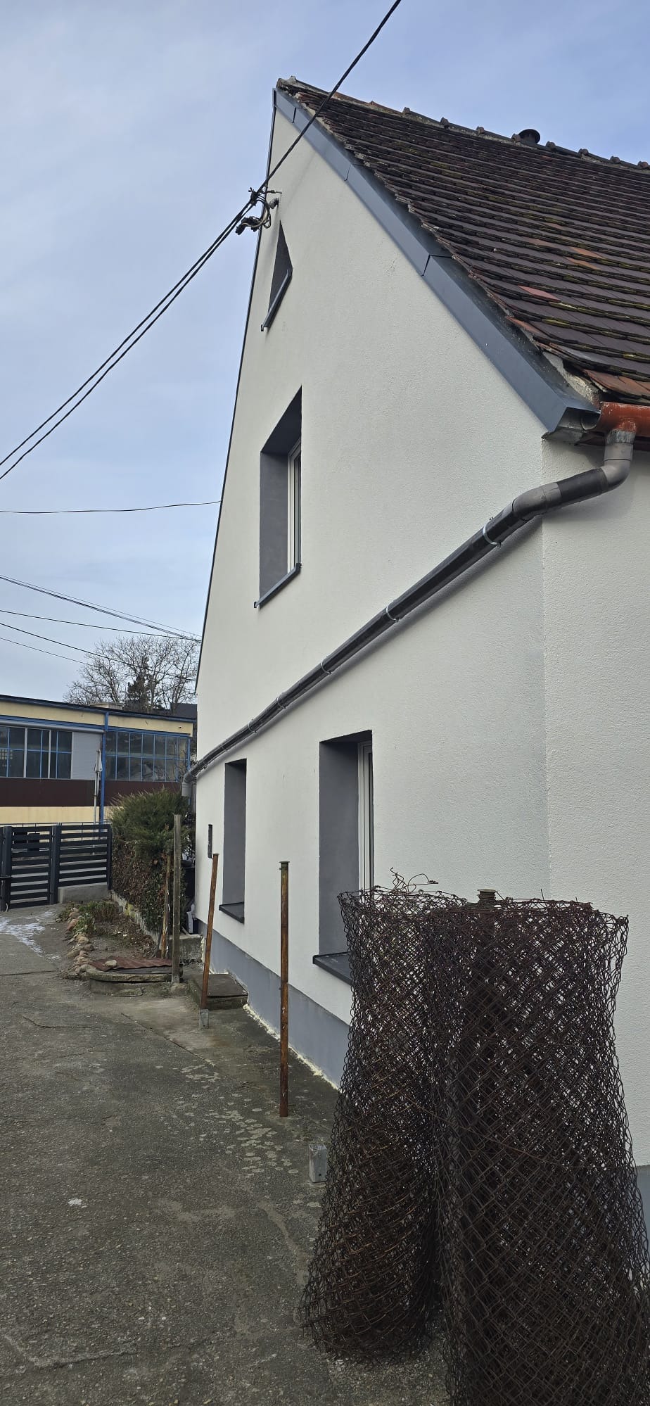 Świeżo otynkowana, biała elewacja domu jednorodzinnego z szarą stolarką okienną i rynną w Opolu. Widok z boku, z fragmentem ogrodzenia z siatki na pierwszym planie.