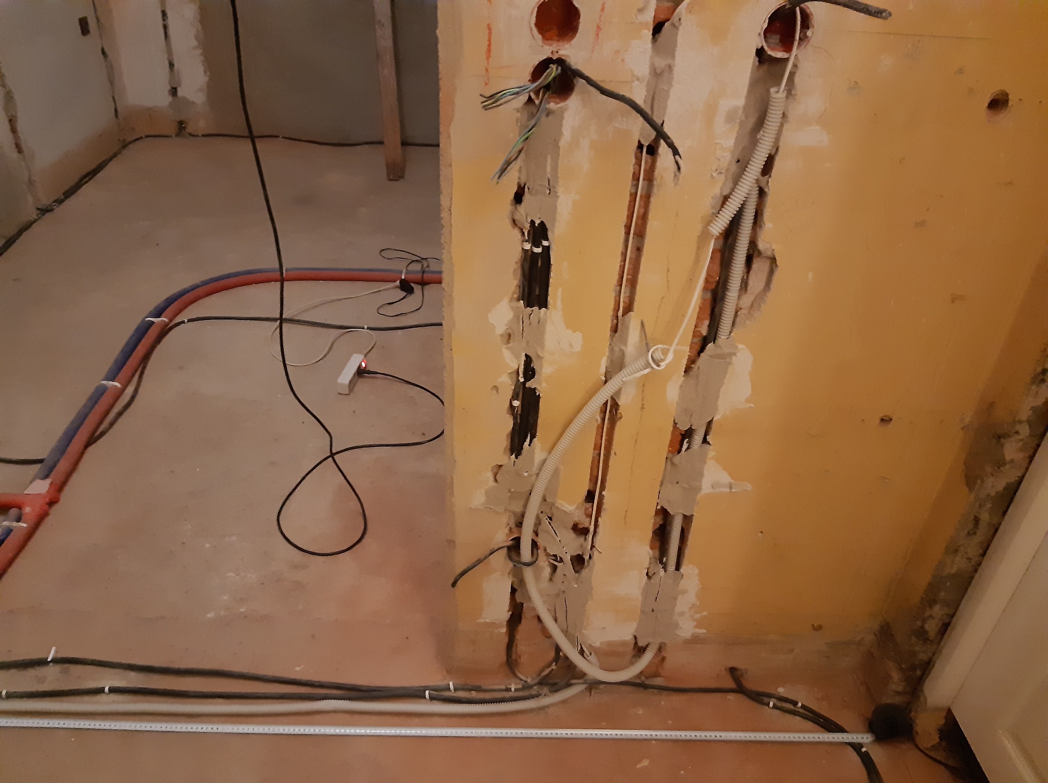 Ściana z bruzdami pod instalacje elektryczne, widoczne kable i rury. Na podłodze leżą przewody i miarka. Prace instalacyjne w trakcie remontu.