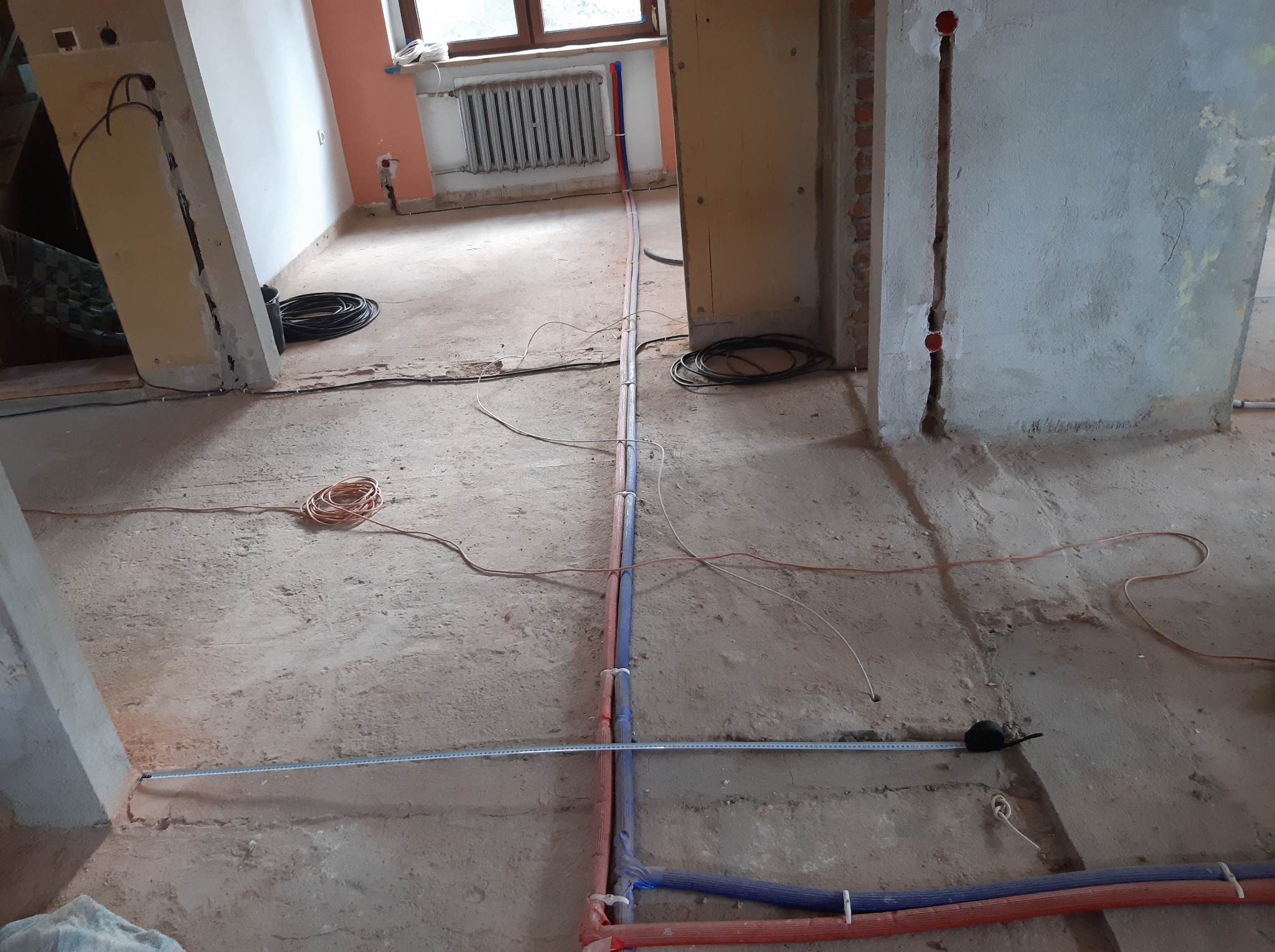 Instalacja rur wodnych i przewodów elektrycznych w surowym wnętrzu. Widoczne bruzdy w ścianach i podłodze, zwinięte kable i miara zwinięta na podłodze.