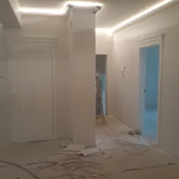 Białe wnętrze w trakcie prac wykończeniowych: świeżo pomalowane ściany, listwy LED pod sufitem, zabezpieczona podłoga, drabina w tle. Widok na dwa wejścia.