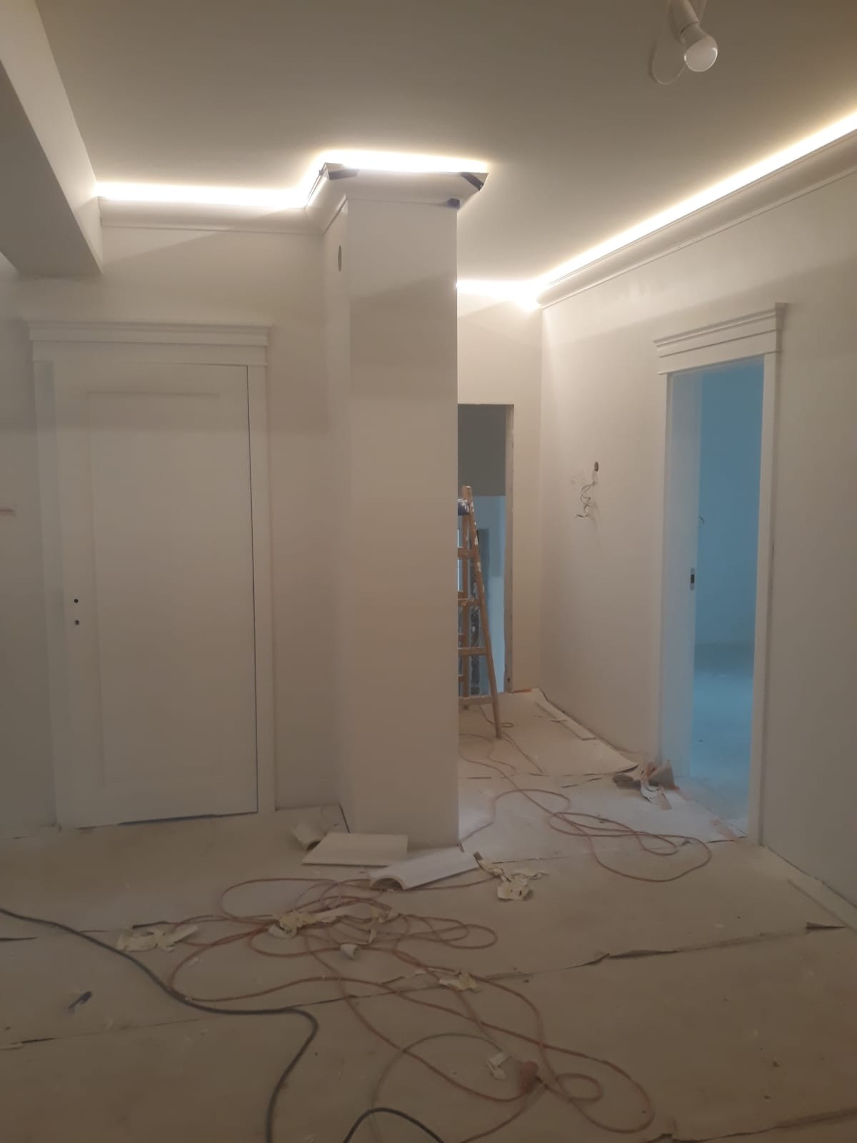 Białe wnętrze w trakcie prac wykończeniowych: świeżo pomalowane ściany, listwy LED pod sufitem, zabezpieczona podłoga, drabina w tle. Widok na dwa wejścia.