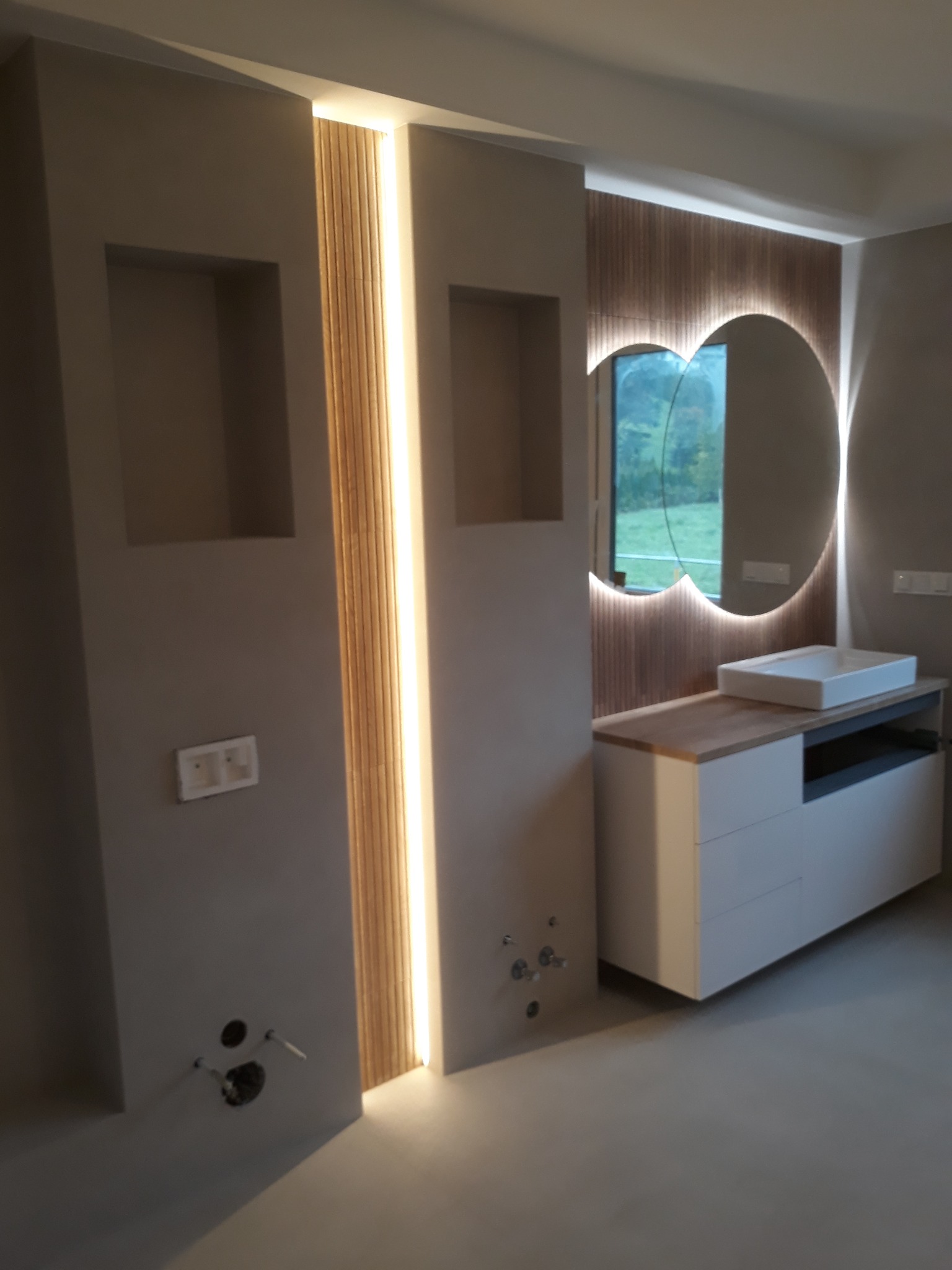 Nowoczesna łazienka w odcieniach szarości z podświetlanym lustrem o nieregularnym kształcie i drewnianymi akcentami. Minimalistyczna umywalka nablatowa.